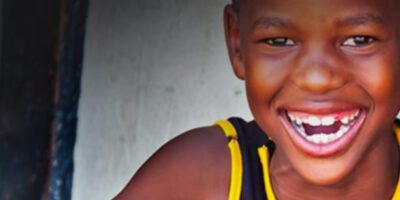 A Young Black Boy, Wearing A Navy Blue Tank Top With Yellow Striping, Smiles Directly Into The Camera With A Big Toothy Grin.