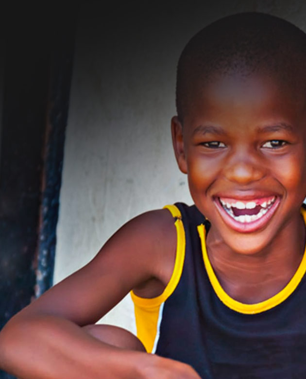 A young Black boy, wearing a navy blue tank top with yellow striping, smiles directly into the camera with a big toothy grin.