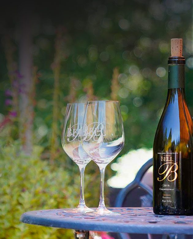 Two long-stemmed wine glasses, bearing an embossed version of the the Bell Wines logo, sit on iron table next to a corked bottle of Bell Wine in an outdoor setting