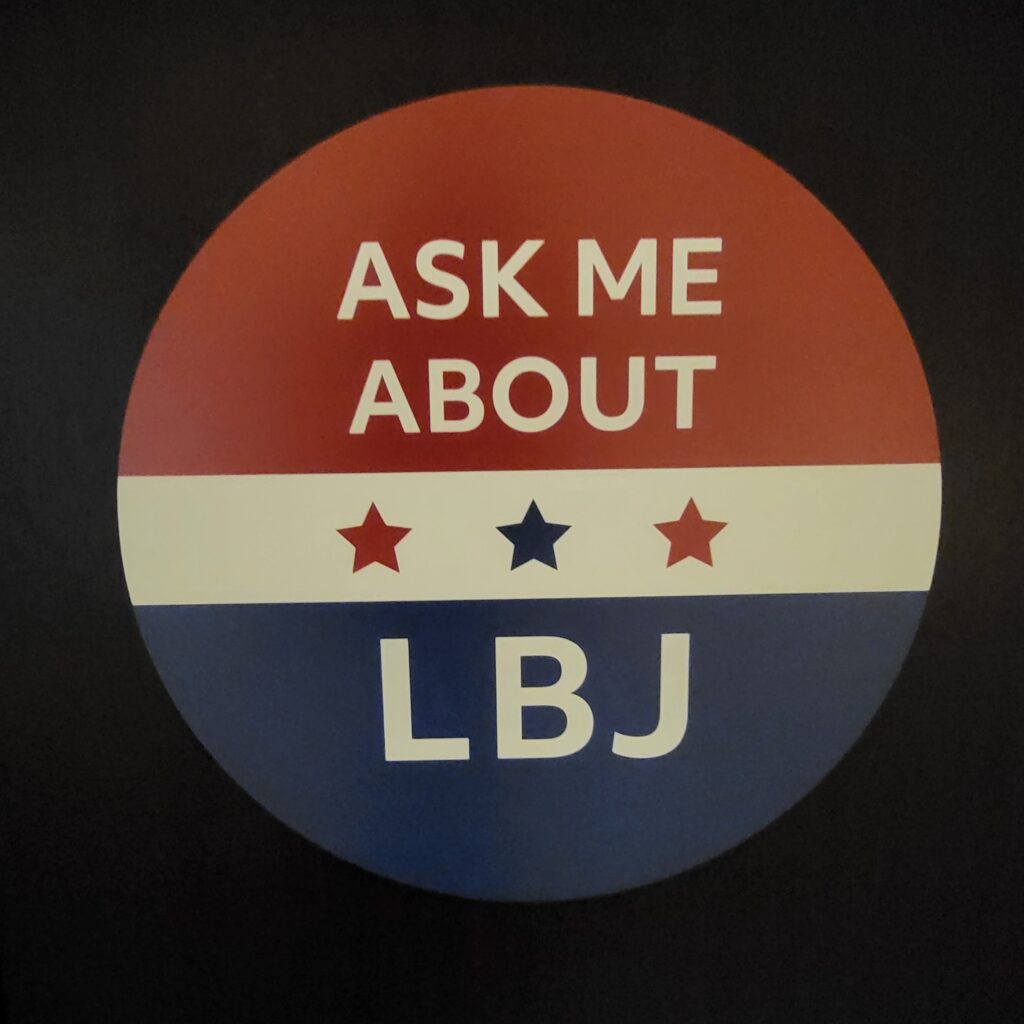 Photo of an red, white, and blue button that reads "Ask me about LBJ" as displayed at the LBJ Presidential Library in Austin, Texas.