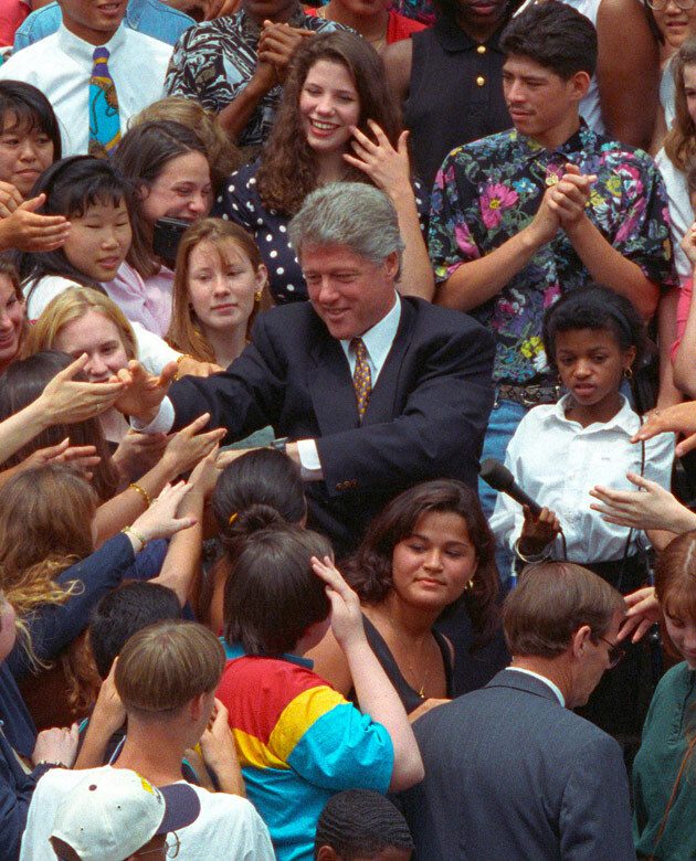 An image of President Bill Clinton in the middle of a crowd of diverse individuals, shaking outstretched hands